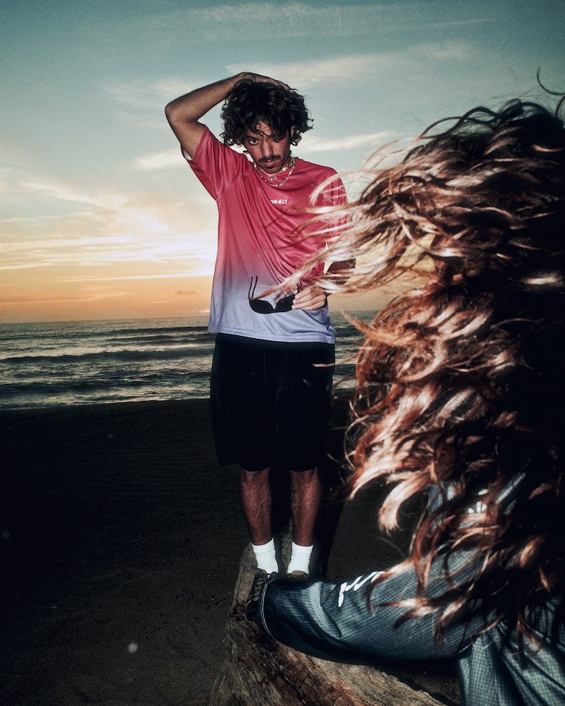 Person standing on a beach with a sunset, wearing a red shirt and black shorts.
