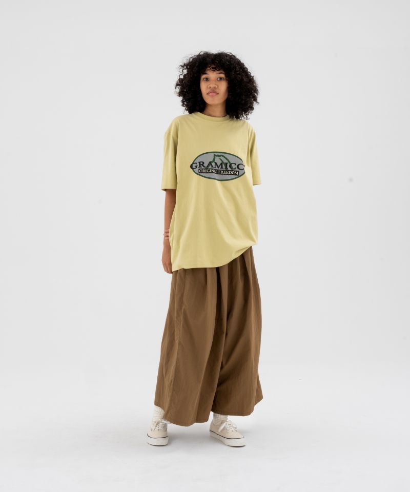Model wearing a foggy lime Gramicci Half Dome Tee with grey graphic and brown pants on a light-tone background.