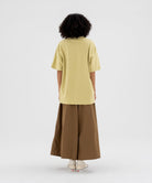 Back view of a model wearing a foggy lime Gramicci Half Dome Tee and brown pants on a light-tone background.