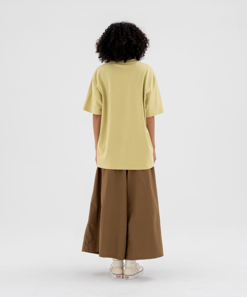 Back view of a model wearing a foggy lime Gramicci Half Dome Tee and brown pants on a light-tone background.