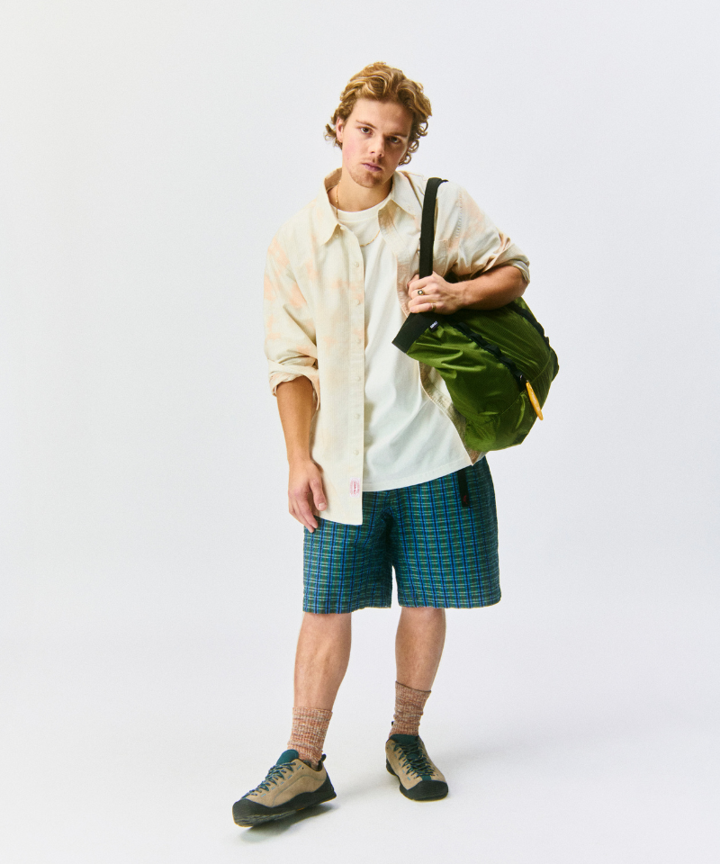 Model wearing a peach moddled shirt, blue plaid shorts, and carrying a green bag on a light-tone background.