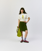 Model wearing a white t-shirt with a Gramicci beverage graphic, green skort,  Hemp Blend Socks, and carrying a green bag on a light-tone background