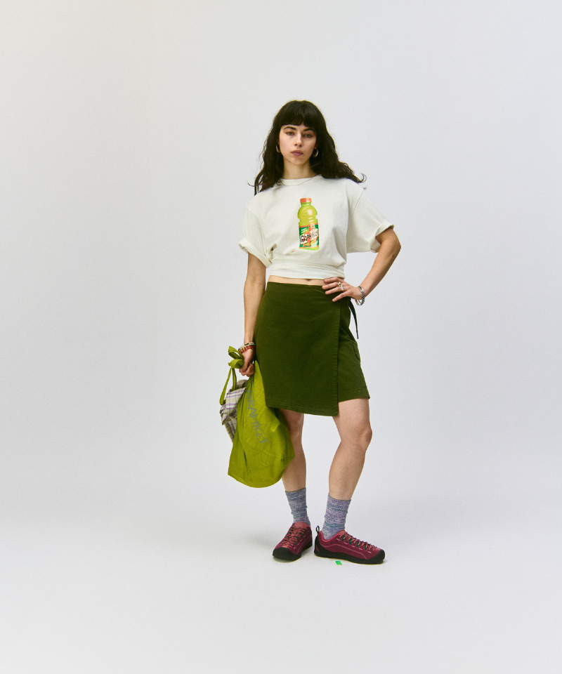 Model wearing a white t-shirt with a Gramicci beverage graphic, green skort,  Hemp Blend Socks, and carrying a green bag on a light-tone background