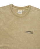 Close-up of a beige mineral-wash t-shirt with a small black embroidered logo on the left chest reading 'GRAMICCI thisisneverthat'.