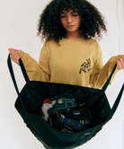 Model holding a green Gamicci Trail Tote and a yellow shirt on a light-tone background.